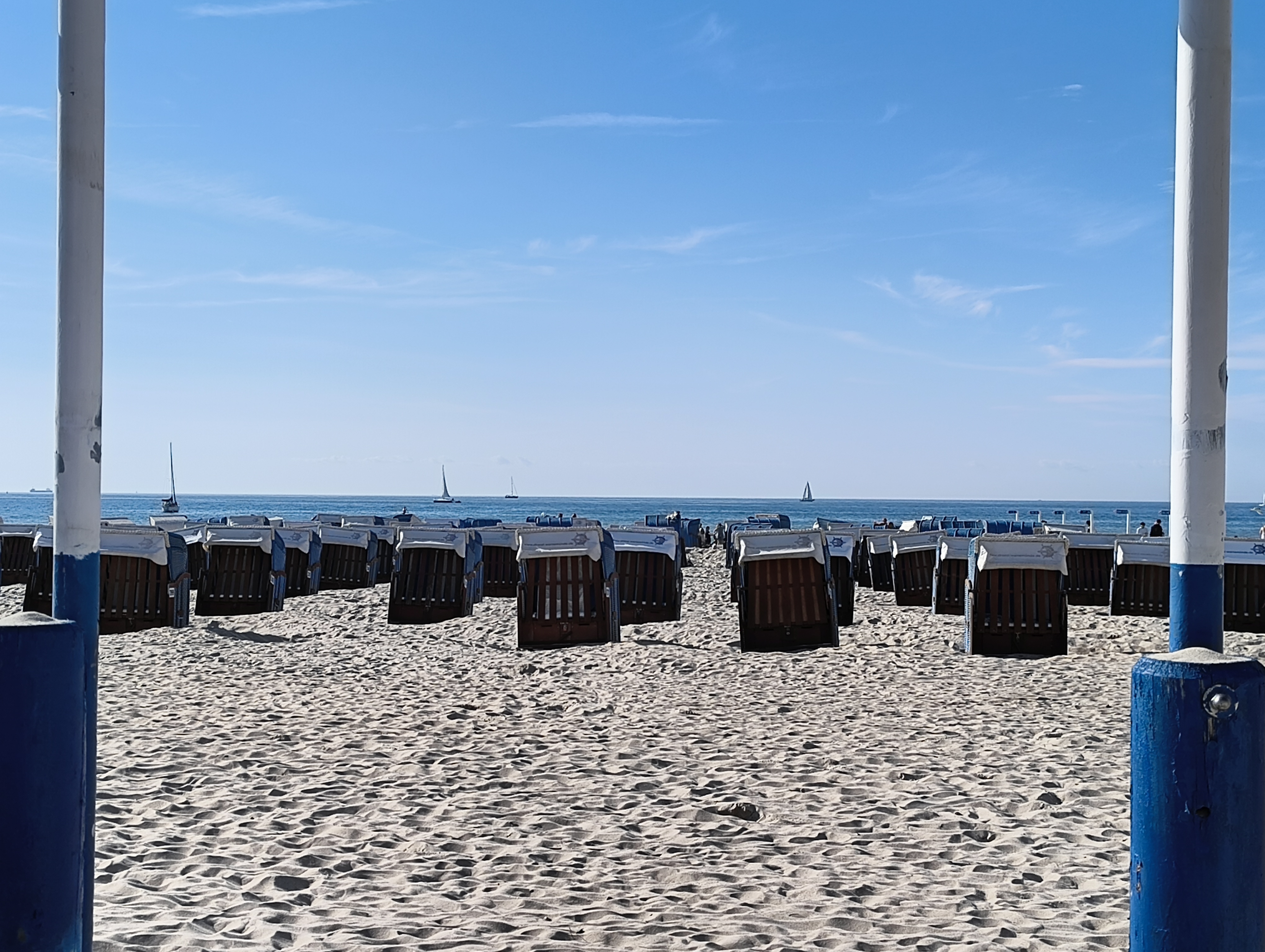 Blick durch zwei Pfosten auf den Strand mit Strandkörben, die hinter- und nebeneinander in Reihen stehen; dabei sind die meisten auf das Wasser ausgerichtet. Im Hintergrund Boote und Schiffe auf dem Wasser in unterschiedlicher Entfernung vom Strand. Der Himmel ist strahlend blau.