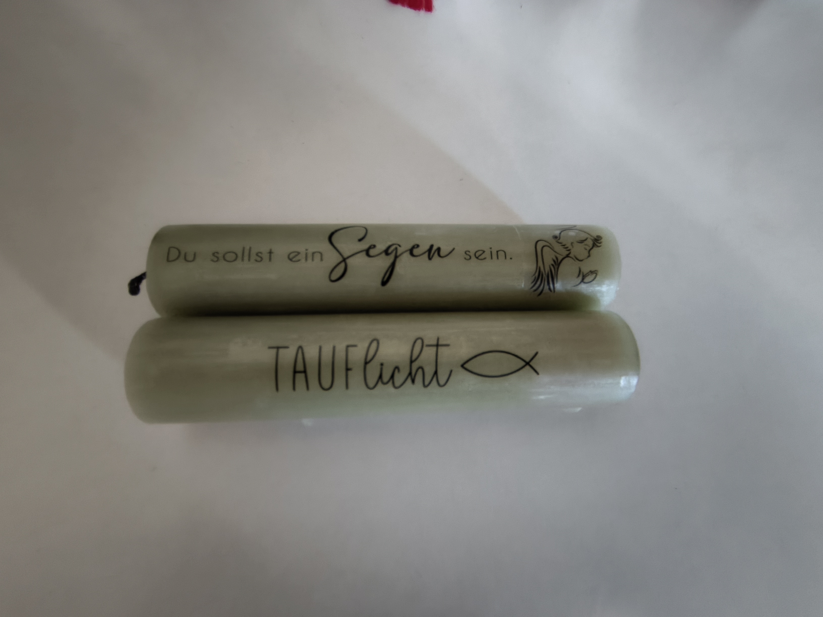 Zwei gleichfarbige Kerzen liegen übereinander auf einer weißen Unterlagen. Auf der oberen Kerze steht der Spruch „Du sollst ein Segen sein“, daneben ein Engel; auf der unteren Kerze das Wort „Tauflicht“, daneben das Symbol eines Fisches.