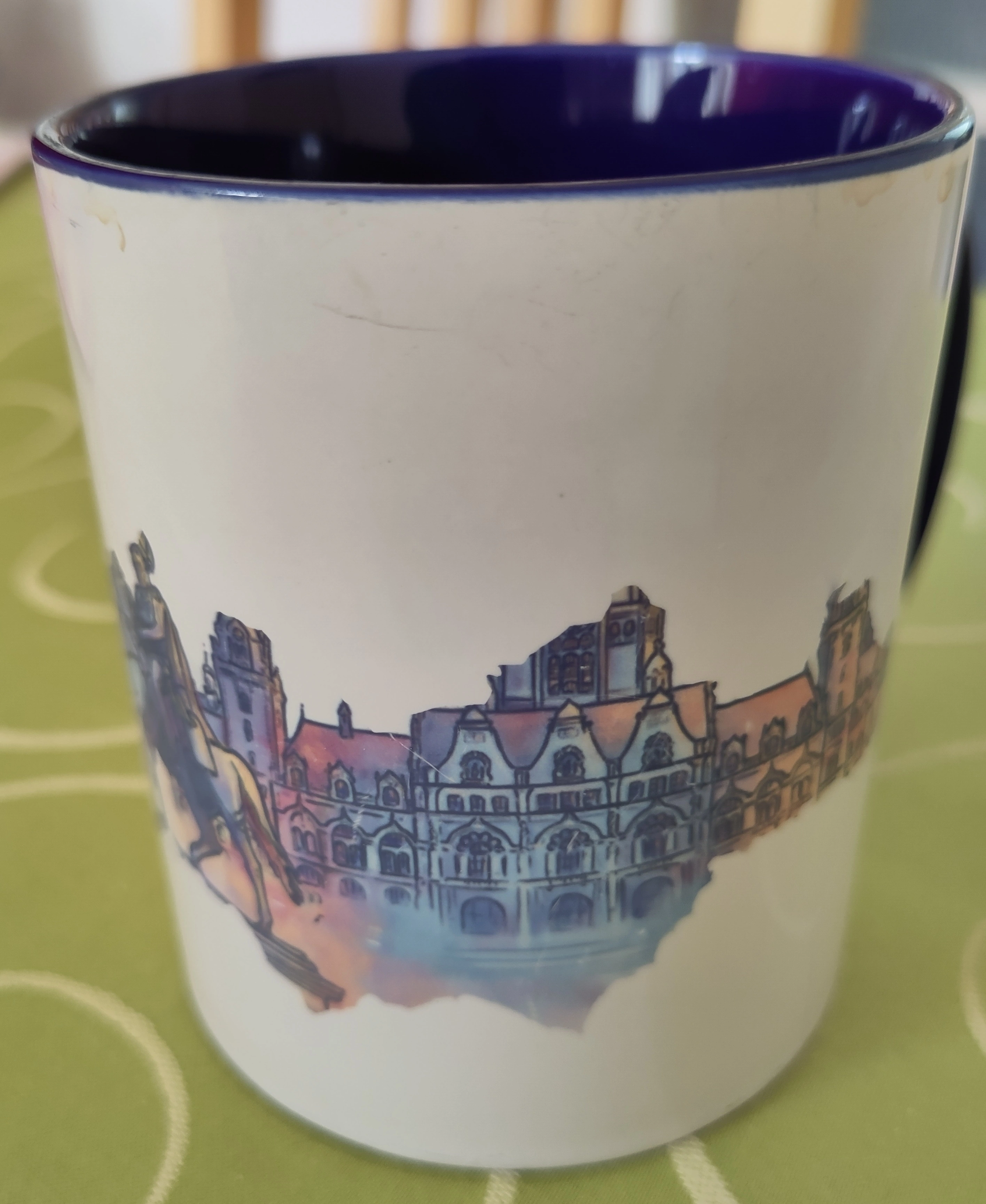Weißer Kaffeebecher mit gemalten Wahrzeichen der Stadt Hannover (unter anderem die Statue vor dem Hauptbahnhof und das Neue Rathaus). Die Innenseite des Kaffeebechers ist dunkelblau. Am Rand der Tasse Kaffeerückstände. Die Tasse steht auf einer grünen Tischdecke mit spiralförmigen Mustern. Im Hintergrund eine Stuhllehne.