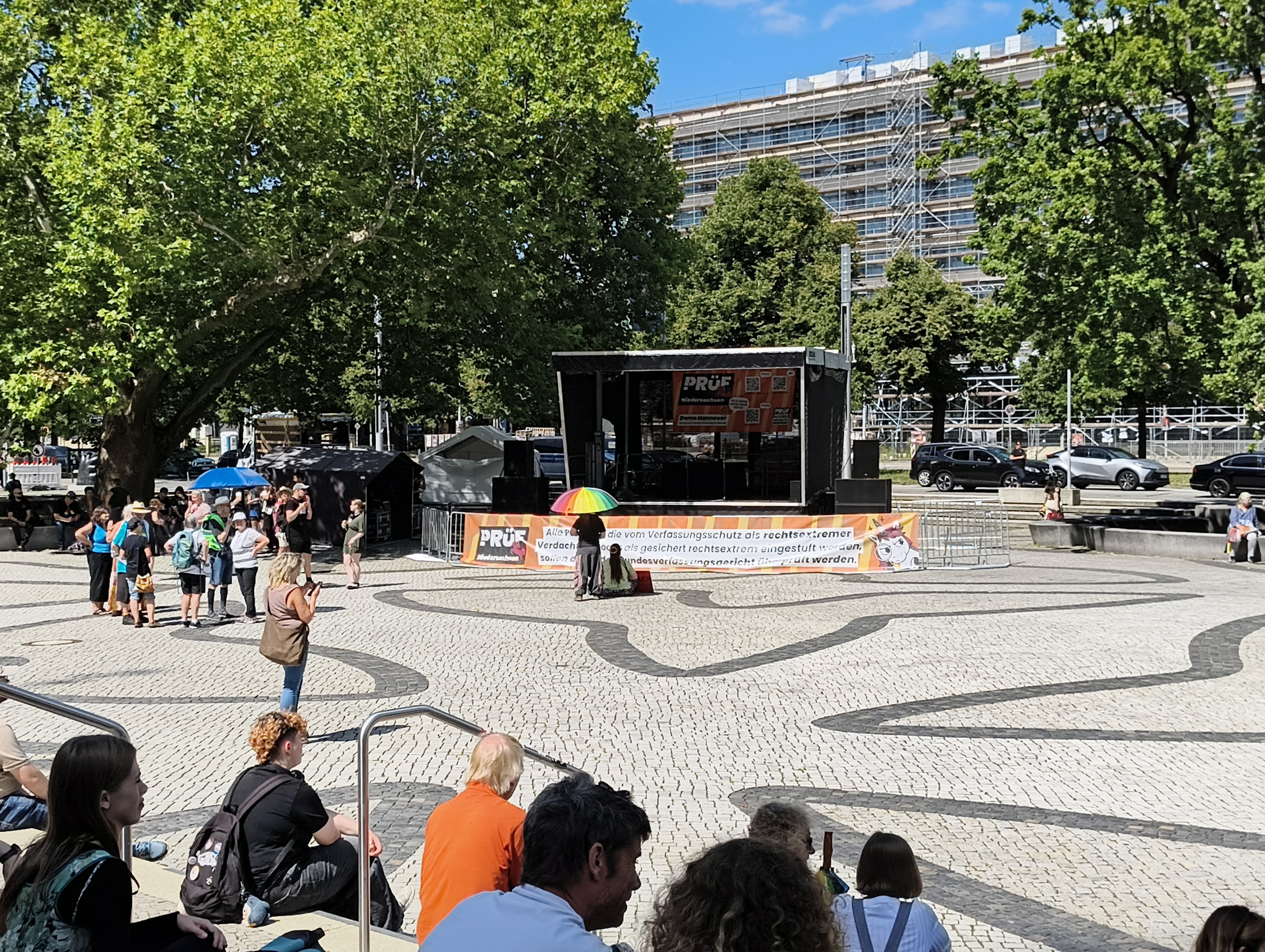 Bühne auf dem Platz der Menschenrechte. Am unteren Ende der Bühne ein breites Banner mit dem Logo von PRÜF Niedersachsen. Nur einige wenige Personen befinden sich direkt auf dem Platz. Markant steht eine Person mit regenbogen-farbenem Regenschirm auf dem Platz und blickt in Richtung Bühne