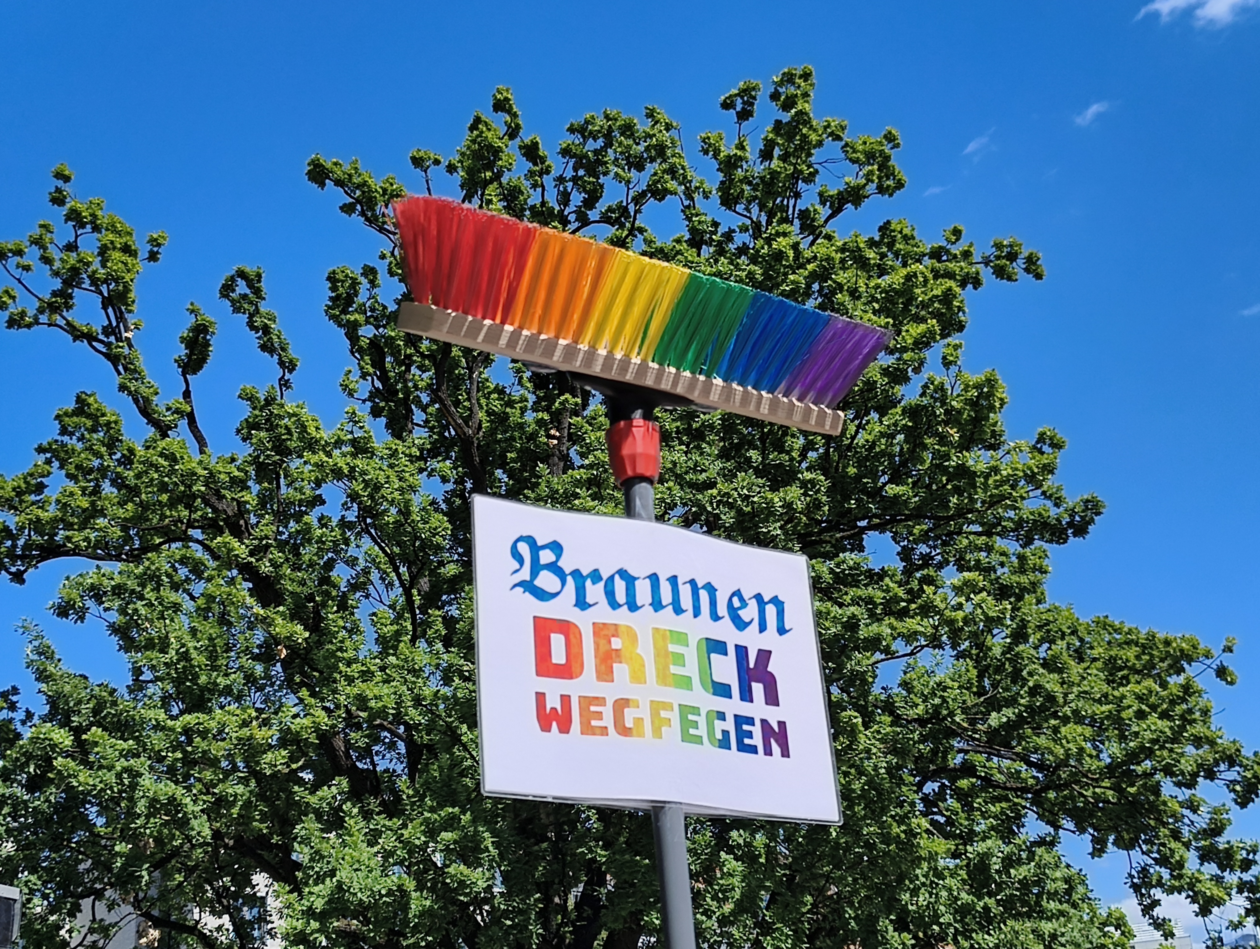 Auf einem Besenstil mit bunten Borsten in Regenbogenfarben ein Schild mit dem Text „Braunen Dreck wegfegen.“