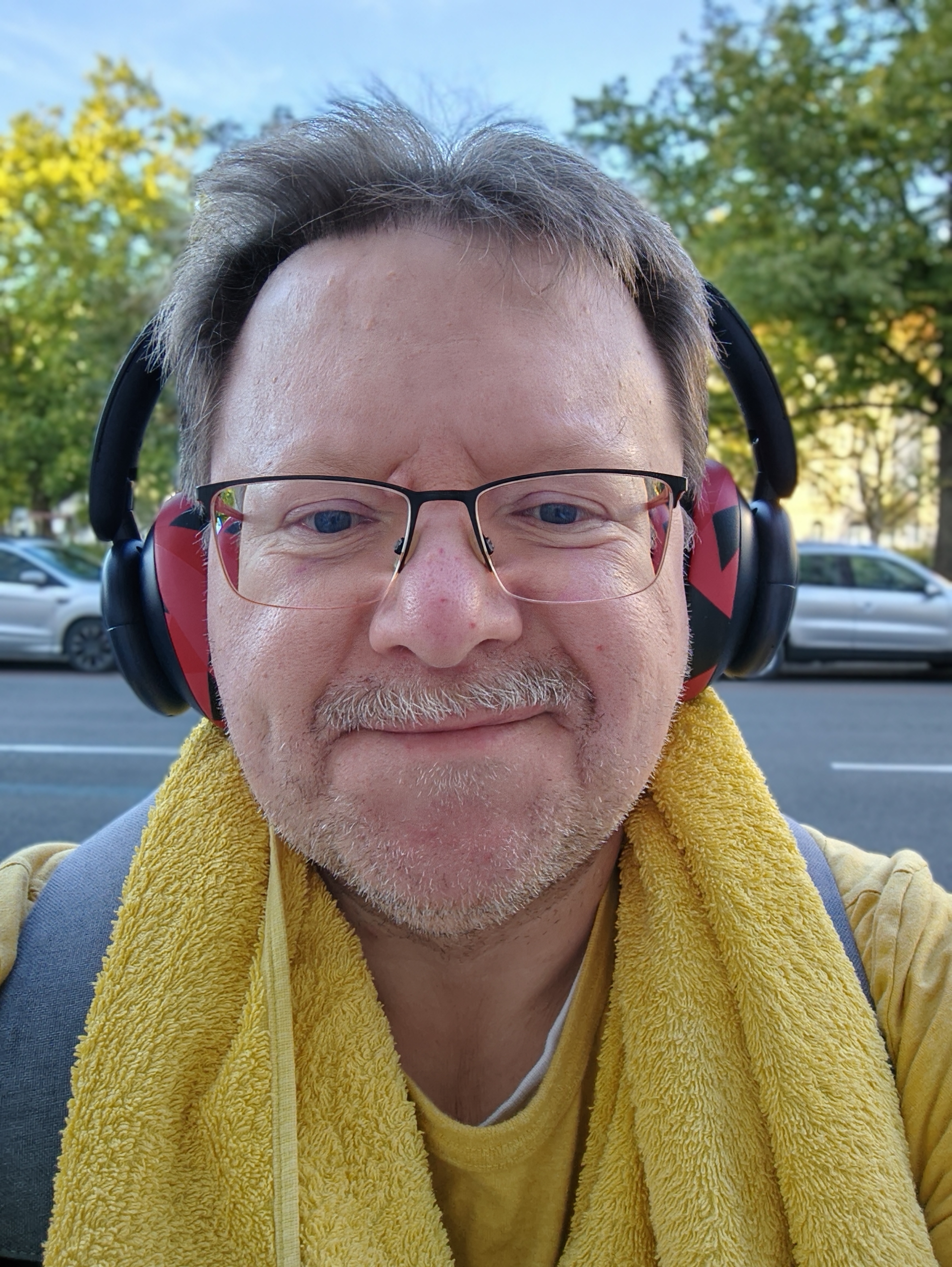 Porträtfoto von mir. Ich trage rot-grau melierte Kopfhörermuscheln an einem dunkelblauen Kopfhörer. Zudem hängt ein gelber Handtuch um meinen Nacken. Die grauen Träger meines Rucksacks sind erkennbar.
