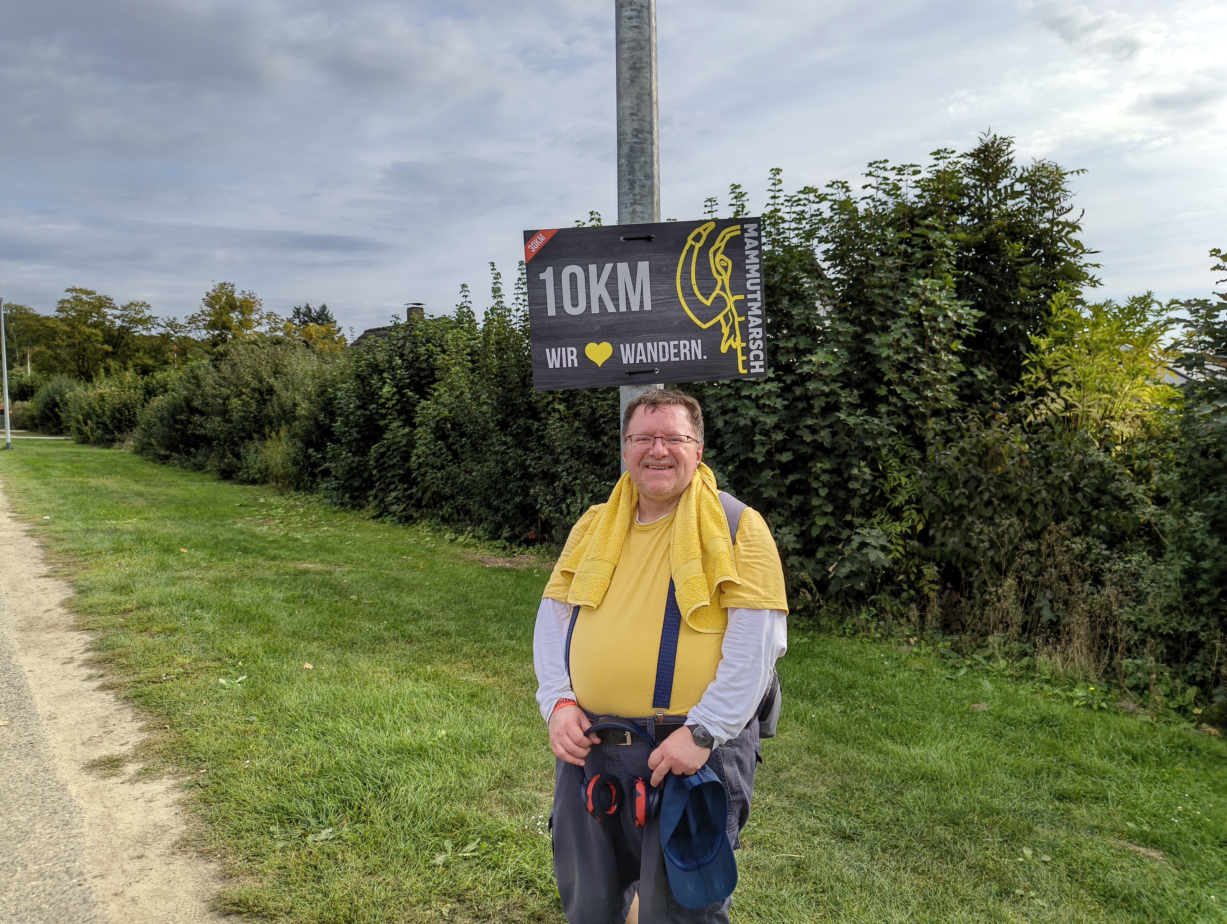 Ich stehe unter dem Schild mit der Aufschrift '10 km · Wir lieben wandern.' Ich trage eine kurze blaue Hose, ein weißen langarmiges T-Shirt, darüber ein gelbes Funktionsshirt. Um den Nacken habe ich ein gelbes Hand liegen. In einer meiner Hände halte ich eine blaue Mütze mit Schirm.