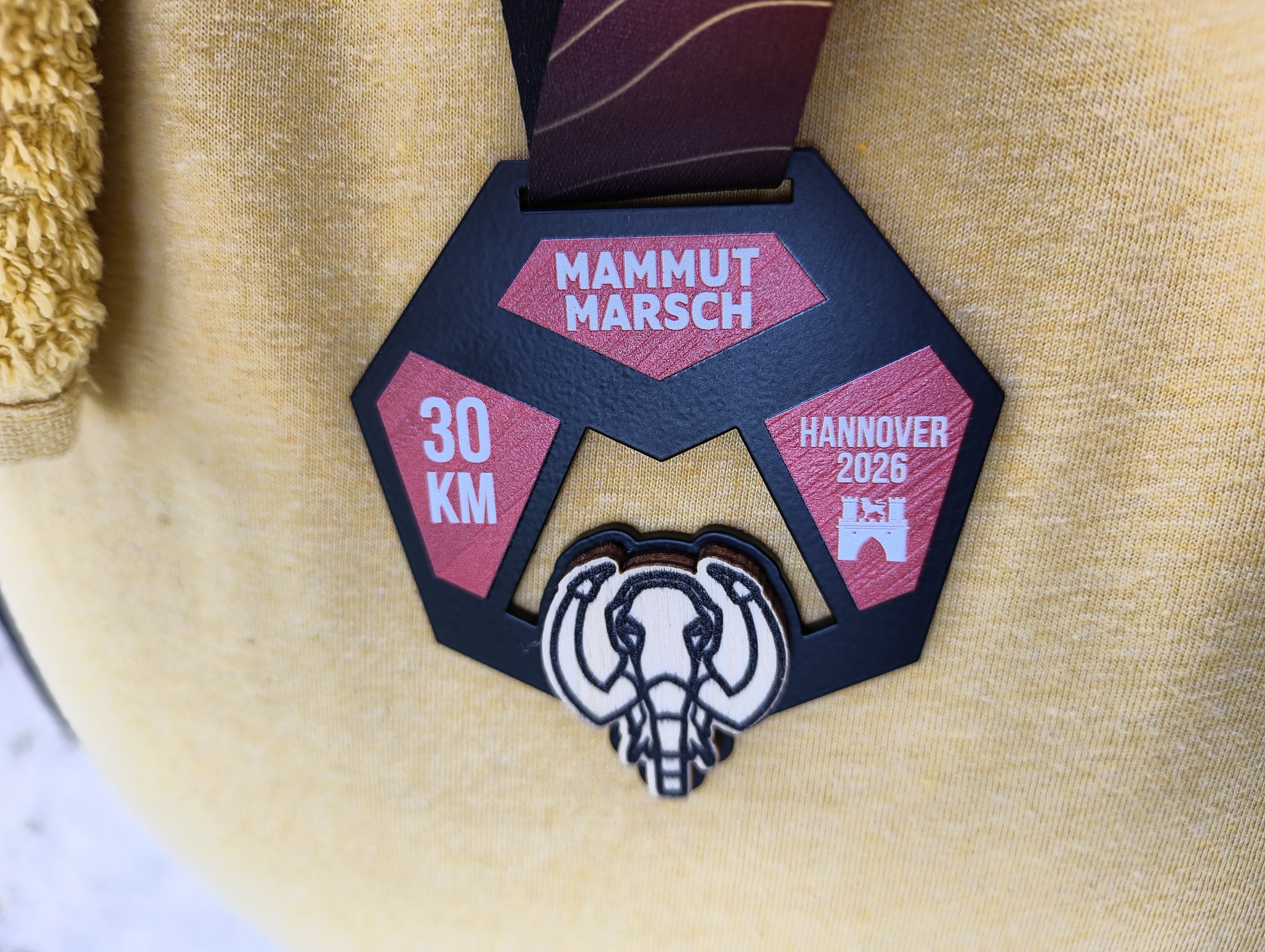 Medaille auf meiner Brust / meinem Bauch auf dem gelbe Funktionsshirt. Unten das Symbol eines Mammuts von vorn. Oben das Wort 'Mammutmarsch', links '30 km' rechts 'Hannover 2026'.