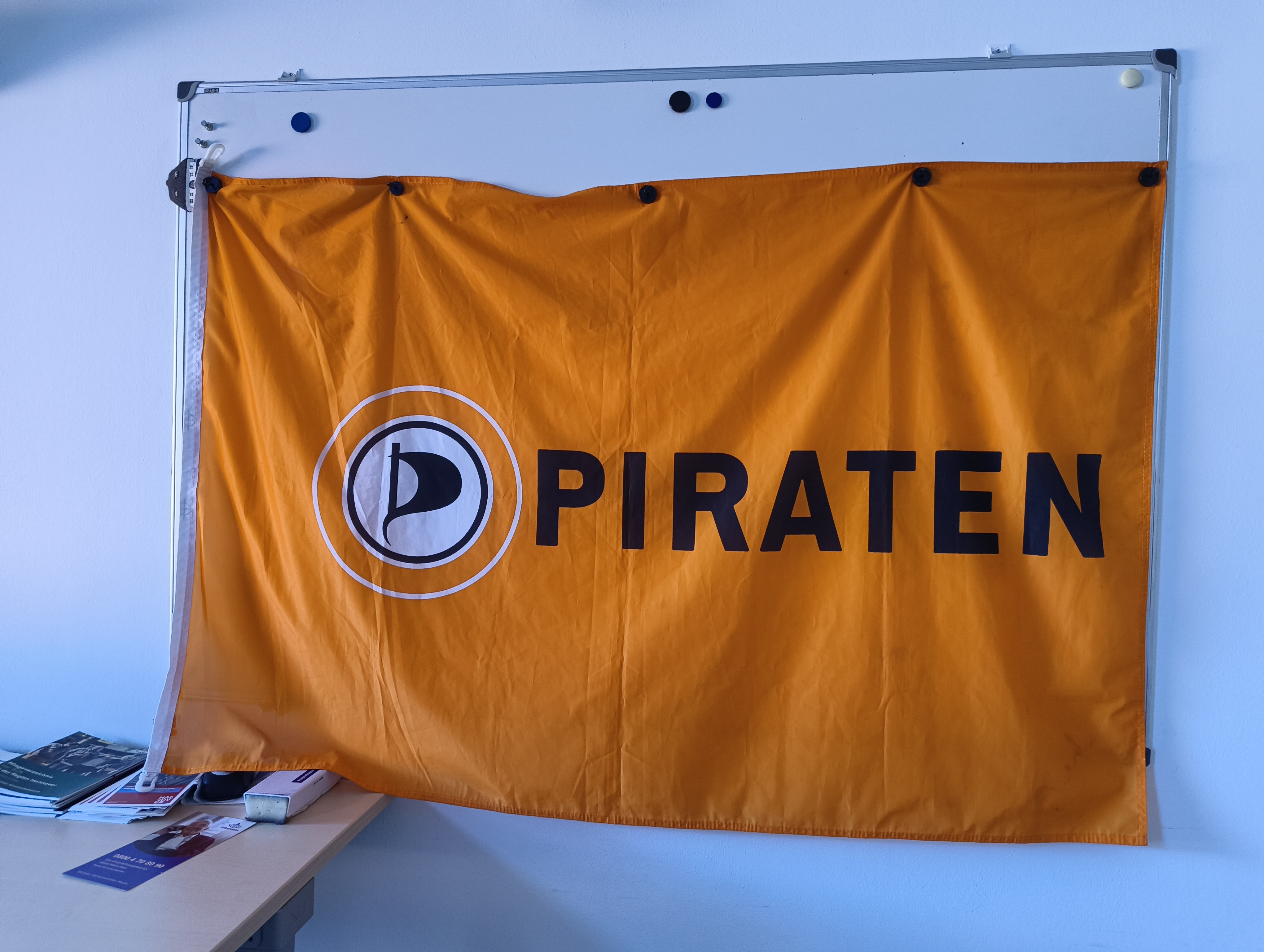 Eine magnetische weiße Tafel an der Wand. Daran wurde mit mehreren schwarzen Magneten eine Piratenflagge befestigt. Seitlich ein großer Arbeitstisch mit Flyern.