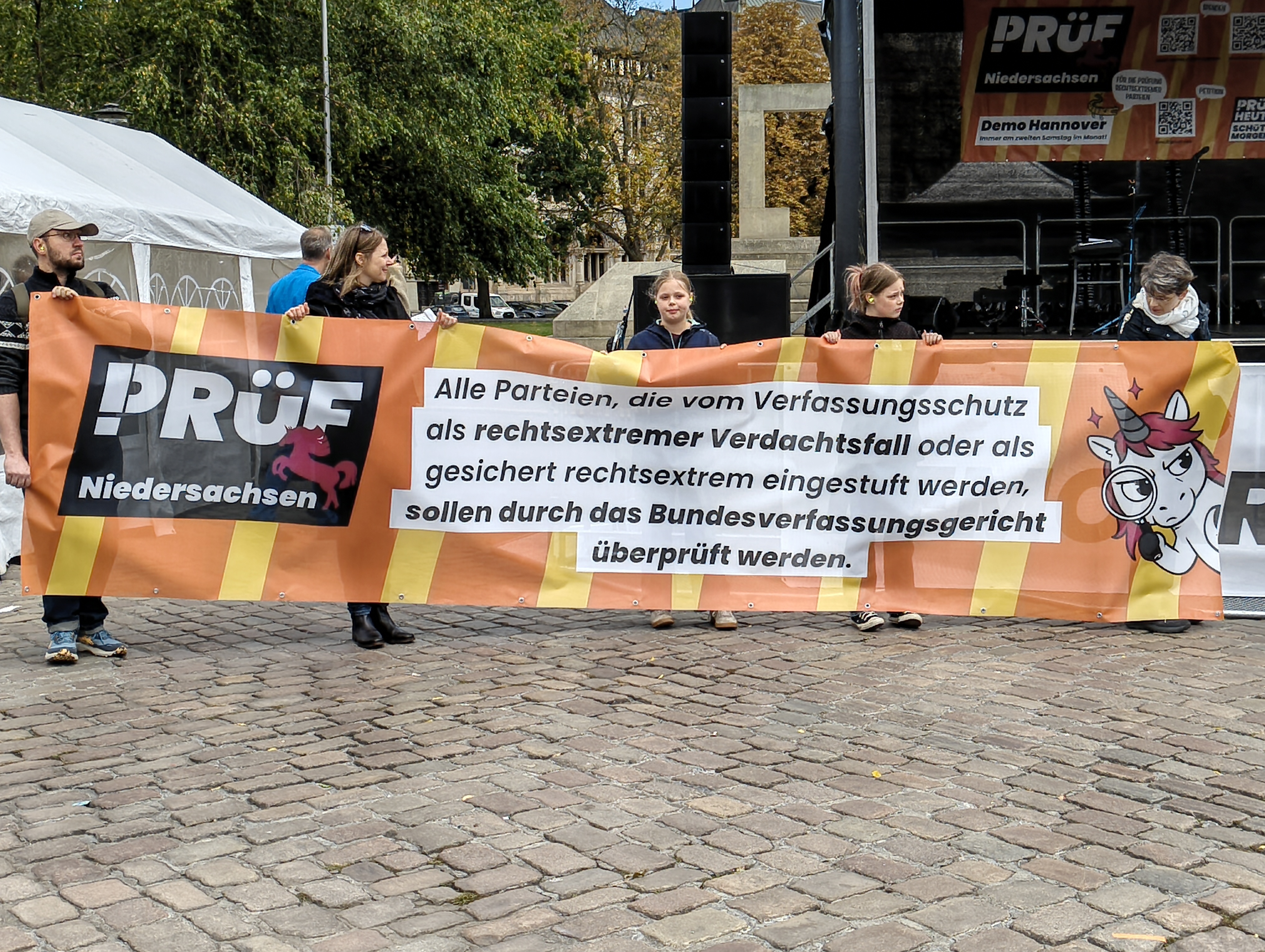 Blick auf einen Bereich vor einer Bühne mit Kopfsteinpflaster. 5 Personen halten ein Banner mit ihren Händen vor ihre Körper. Neben dem Logo PRÜF Niedersachsen steht der Text »Alle Parteien, die vom Verfassungsschutz als rechtsextremer Verdachtsfall oder als gesichert rechtsextrem eingestuft werden, sollen durch das Bundesverfassungsgericht überprüft werden.«