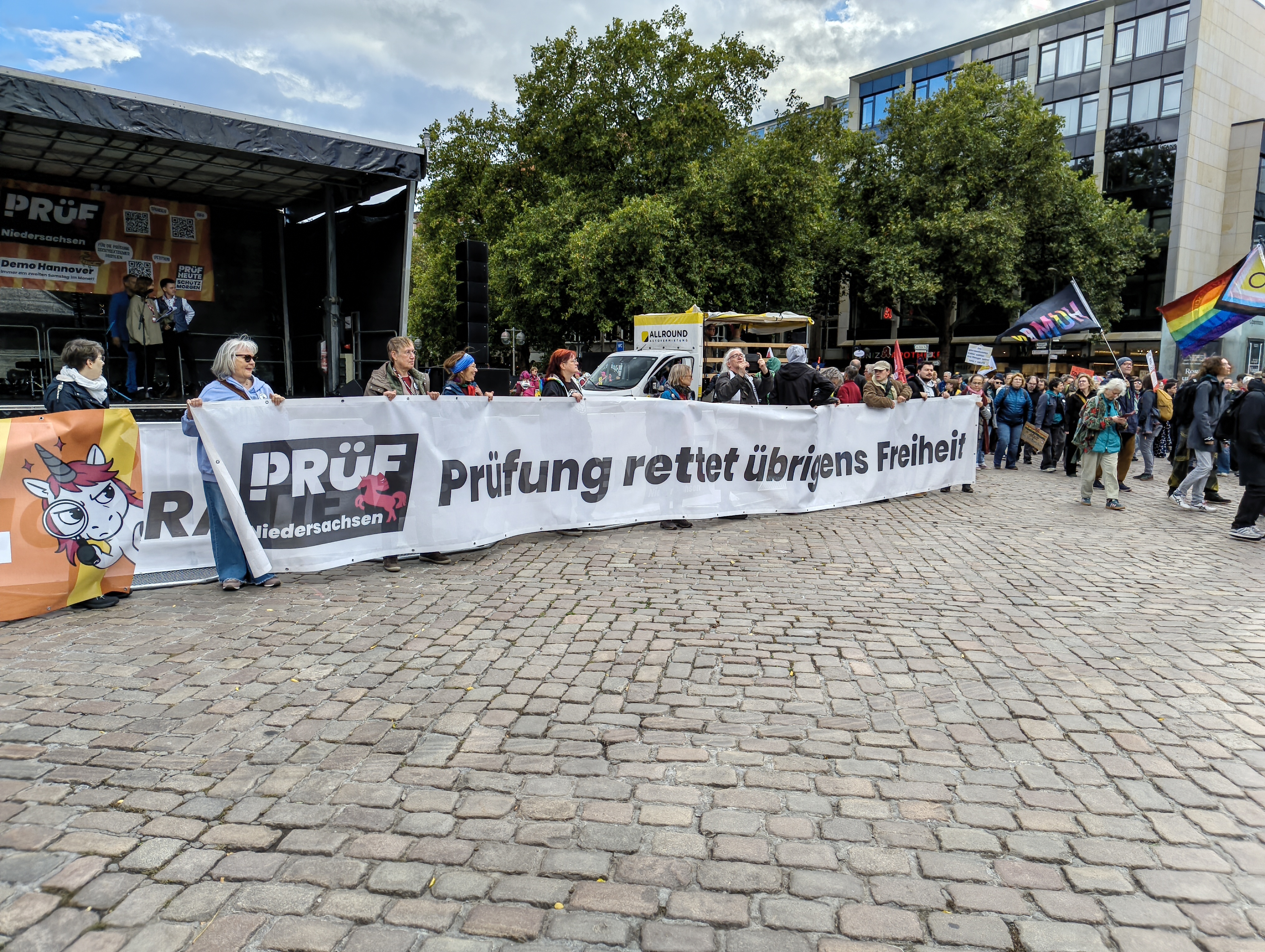 Blick auf einen Bereich vor einer Bühne mit Kopfsteinpflaster. Etwa 10 Personen halten ein Banner mit ihren Händen vor ihre Körper. Auf dem Banner ist neben dem Logo PRÜF Niedersachsen der Text »Prüfung Rettet Übrigens Freiheit« zu lesen. Am rechten Rand (des Bildes) kommen Teilnehmende auf den Platz vor der Bühne.