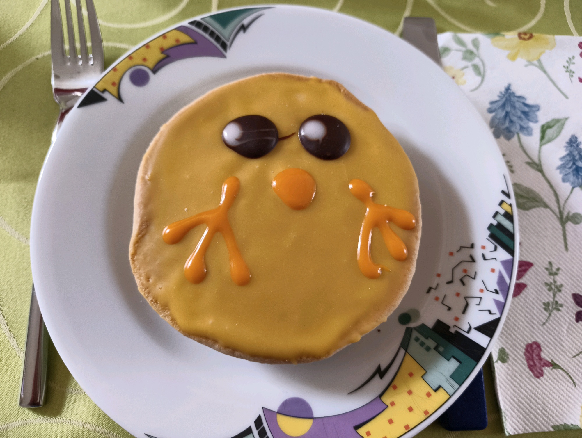 Auf einem runden Kuchenteller liegt ein Gebäckstück, welches Amerikaner genannt wird. Auf dem Zuckerguss das Bild eines Kückens.