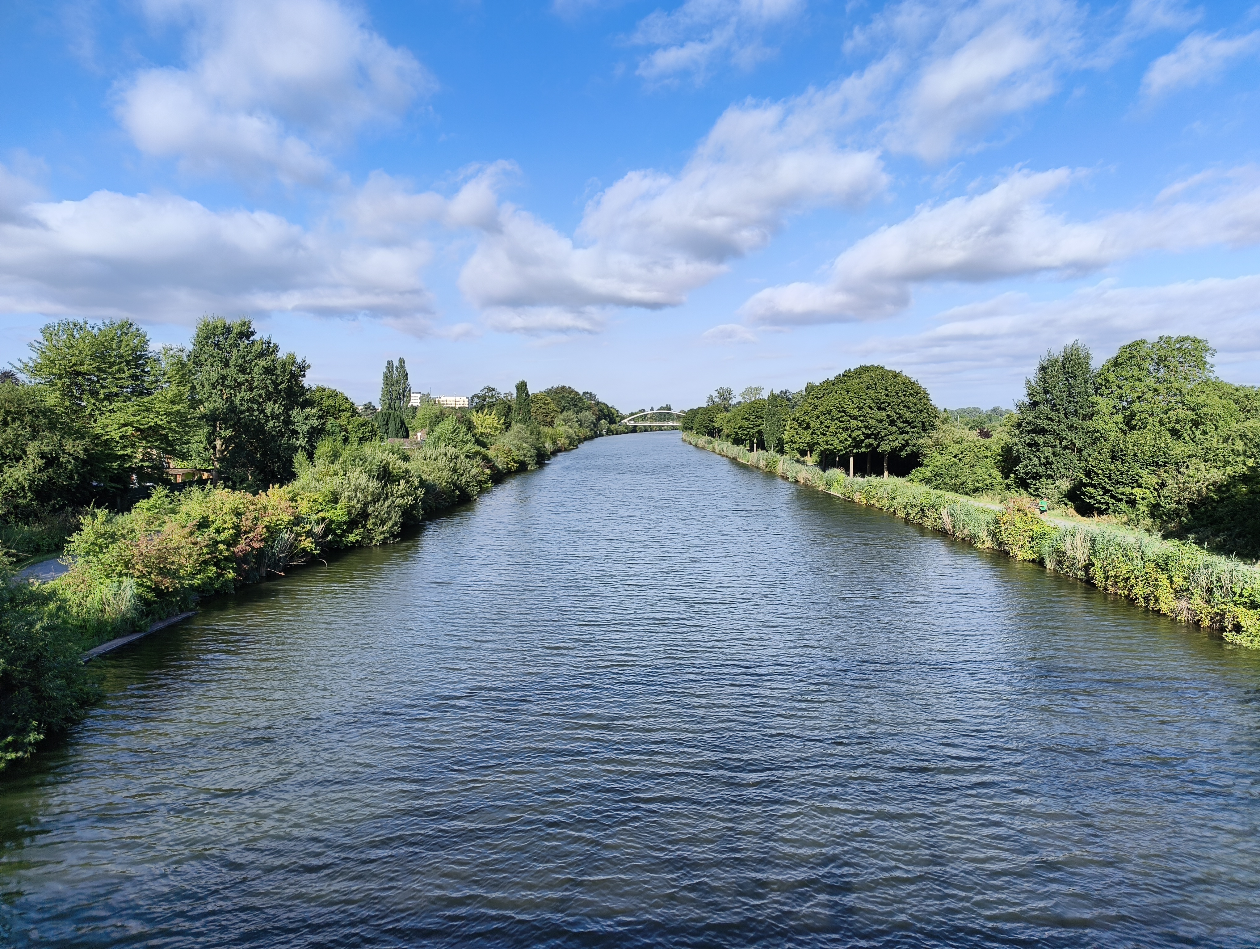 Blick von einer Brücke auf den Mittellandkanal. Rechts und links neben dem Kanal Büsche und Bäume im satten Grün. Im etwas Entfernung eine weitere bogenförmige Brücke. Die stille Wasserfläche spiegelt das helle Tageslicht wider; der strahlend blaue Himmel wird von weißen, lockeren Wolken durchzogen.