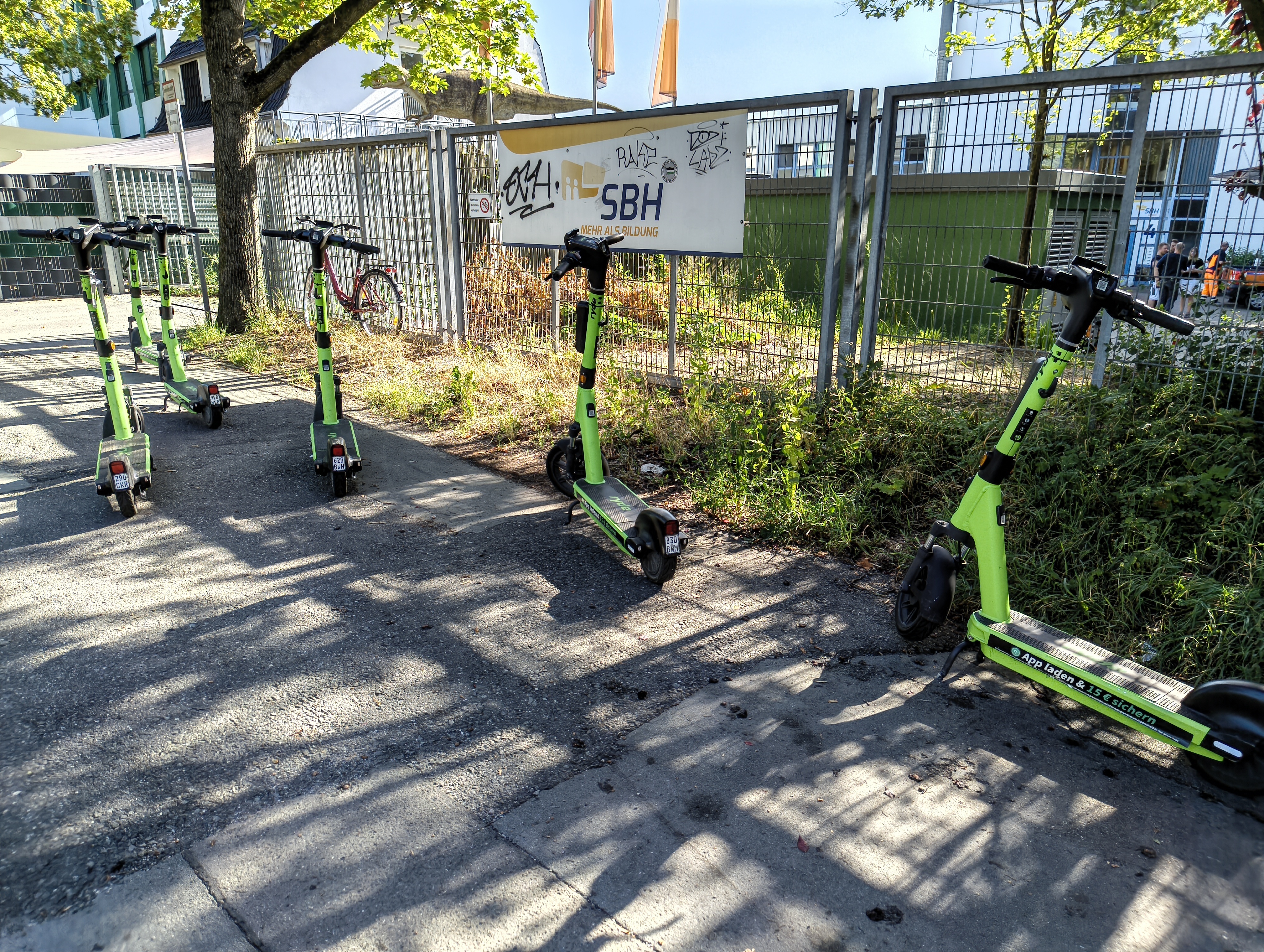 Auf dem Fussweg, in der Nähe eines Zauns stehen 6 grün-weiße eScooter. Auf dem Zaun ein Schild mit der Beschriftung „SBH · Mehr als Ausbildung“. Das Schild ist mit einigen Spray-Namenskürzeln versehen.