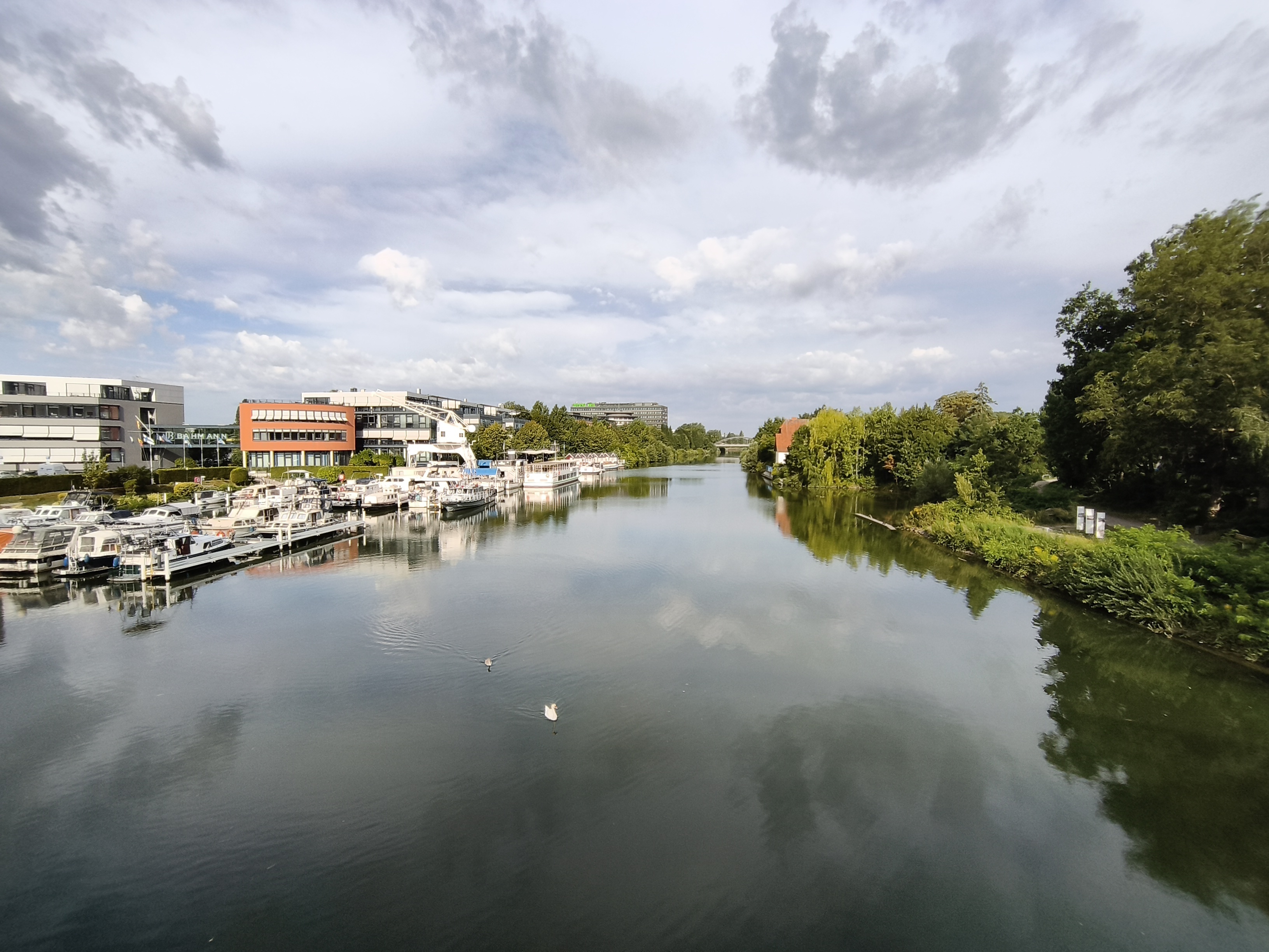 Blick von der Brücke auf beide Seiten des Kanals: Links der Yachthafen mit Schiffen und Booten, rechts Büsche, Bäume und drei weißen Stelen. In Richtung Brücke schwimmen zwei Schwäne.