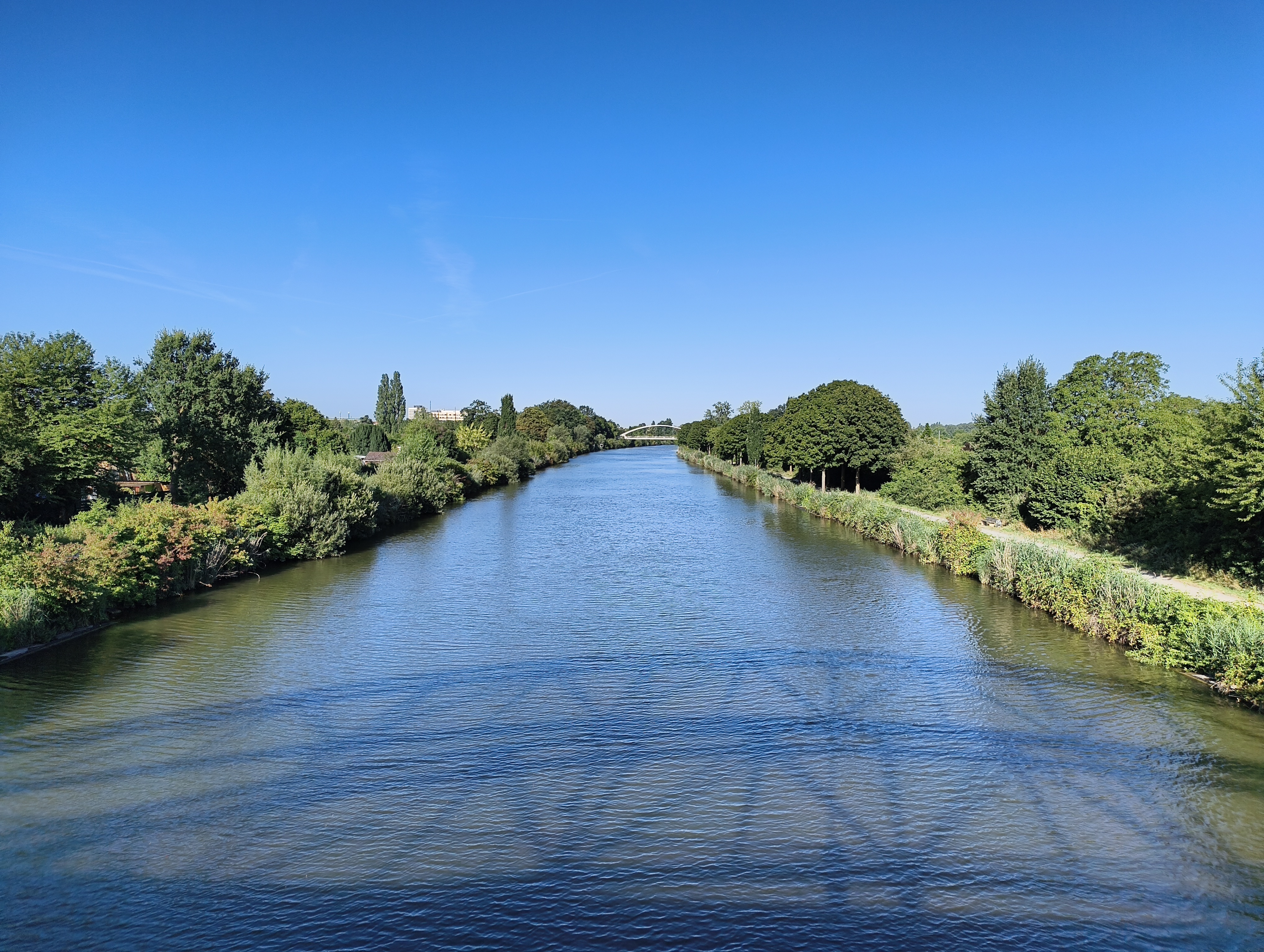 Blick von der Brücke Am Listholze in Richtung Brücke am Lister Damm. Das Wasser im Kanal kräuselt sich nur ganz leicht. Rechts und links sattes Grün von Büschen und Bäumen. Auf der rechten Seite ist der unbefestigte Weg parallel zum Kanal sichtbar. Ein komplett wolkenloser blauer Himmel.