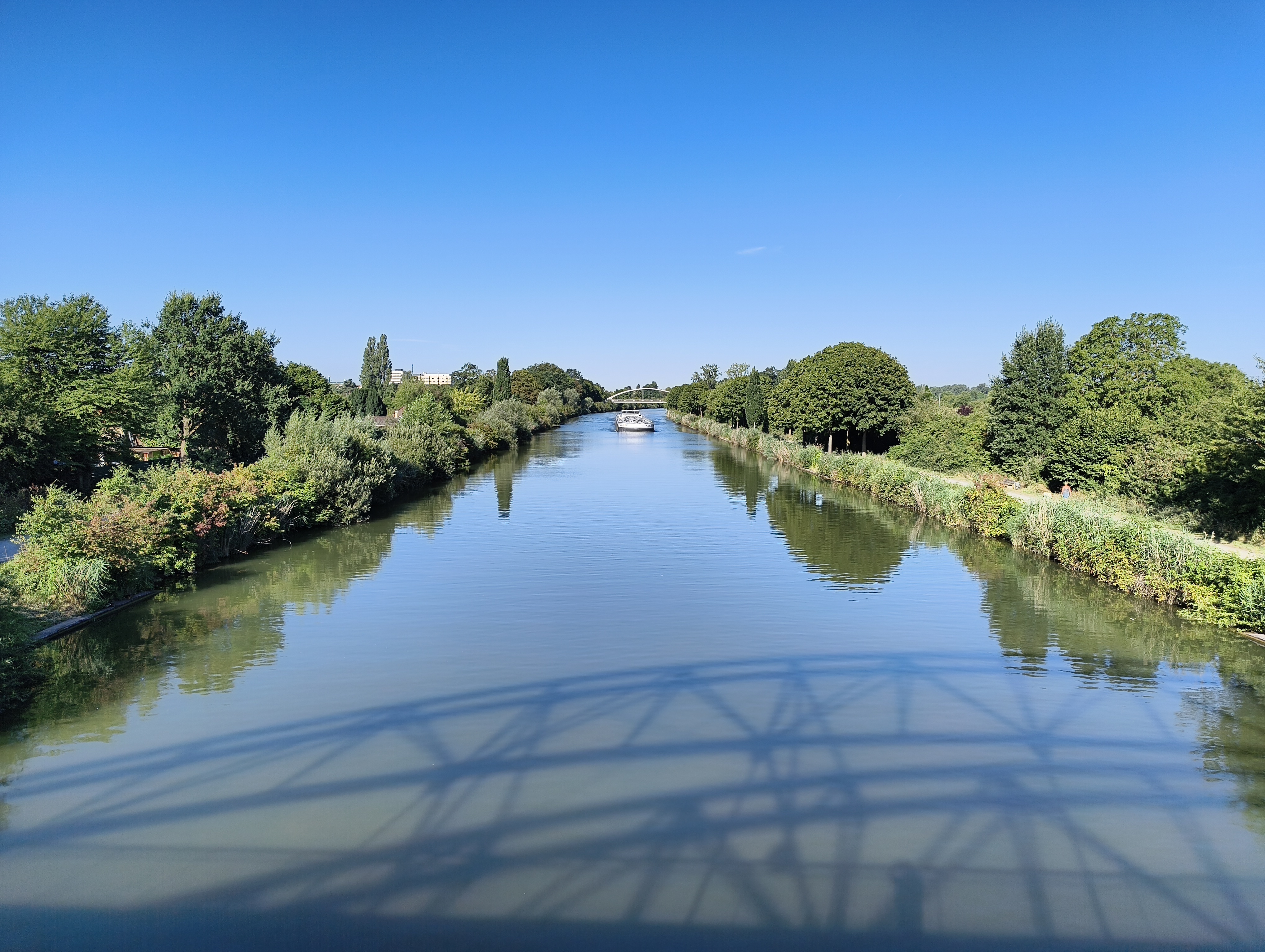 Blick von einer Brücke auf den Mittellandkanal. Rechts und links Büsche und Bäume im satten Grün. Ein Teil des parallel verlaufenden unbefestigten Weges auf der rechten Seite ist deutlich erkennbar. Auf dem Mittellandkanal ein Lastkahn, der auf die betrachtende Person zufährt.